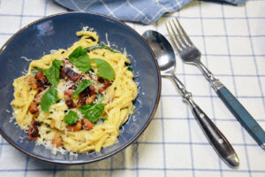 Cremet pastaret med bacon, spinat og revet parmesan i en blå skål på et blåternet viskestykke