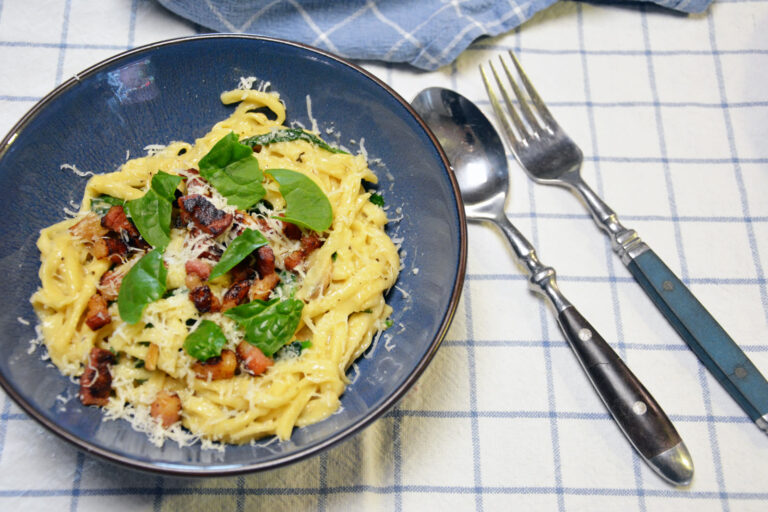Cremet pastaret med bacon, spinat og revet parmesan i en blå skål på et blåternet viskestykke