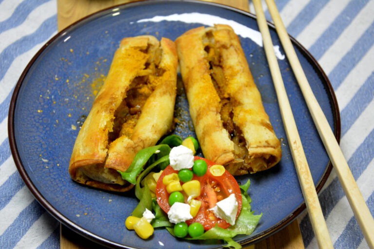 To sprøde forårsruller i airfryer skåret op på en blå tallerken med en lille salat af tomat, majs, ærter og feta og et par spisepinde ved siden af