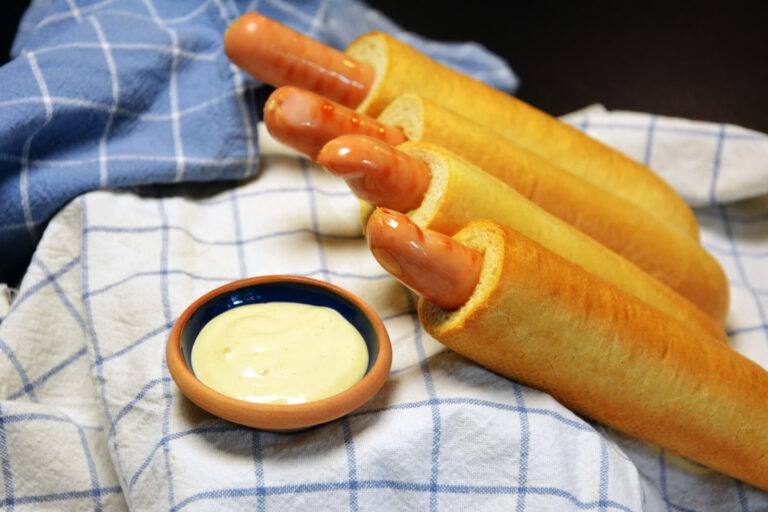 Fire hjemmebagte franske hotdogs med pølse i hullet ligger på et blåternet viskestykke ved siden af en lille skål med lys french dog dressing