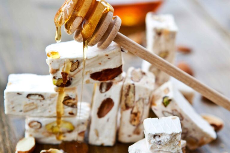 Stykker af hvid fransk nougat med hele mandler og pistacienødder stablet på et træbord, mens honning drypper ned over dem fra en honningske