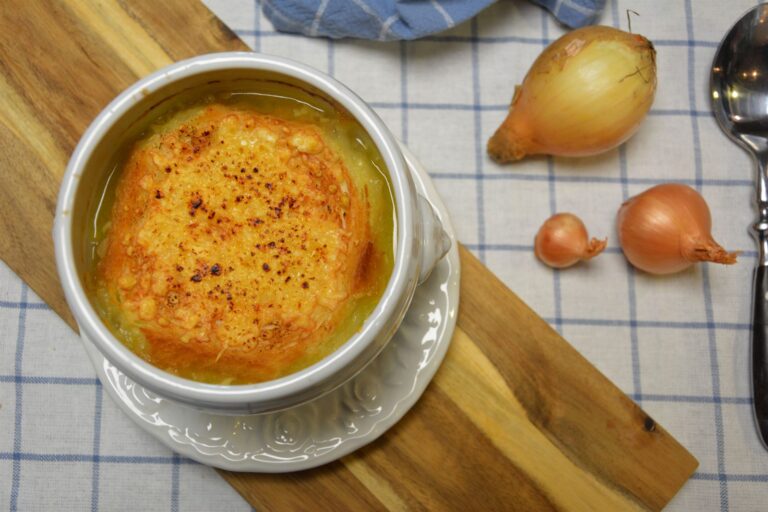 Fransk løgsuppe i en hvid portionsskål med gratineret ostebrød ovenpå, på et træbræt ved siden af hele løg og et blåternet viskestykke