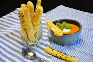 Hjemmebagte grissini med sesamfrø og nigellafrø i et glas ved siden af en skål tomatsuppe
