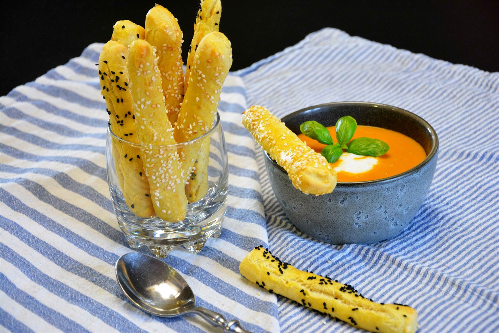 Hjemmebagte grissini med sesamfrø og nigellafrø i et glas ved siden af en skål tomatsuppe