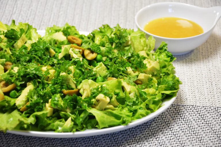 Grøn salat med krølsalat, avocado, grønne oliven, ærter og persille på et hvidt fad med vinaigretten i en skål ved siden af