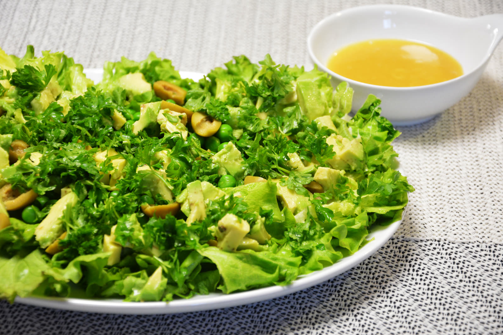 Grøn salat med krølsalat, avocado, grønne oliven, ærter og persille på et hvidt fad med vinaigretten i en skål ved siden af