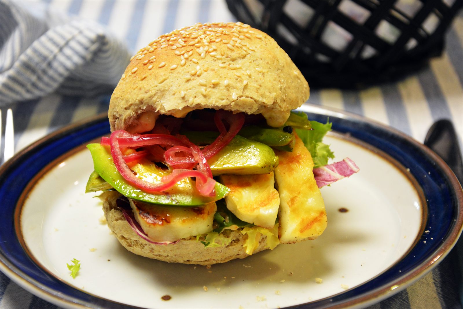 Halloumi-burger i sesambolle med grillstribede halloumiskiver, avocado, syltede rødløg og salat på en tallerken med blå kant