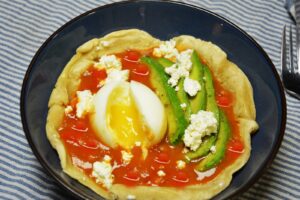 Huevos rancheros i en dyb blå skål: en blød hvedetortilla med salsa roja, et halveret smilende æg, avocadoskiver og smuldret salatost