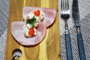 Italiensk salat med gulerodstern og ærter lagt oven på to foldede skiver kogt skinke på rugbrød, pyntet med en halv cherrytomat og dild, på et træskærebræt