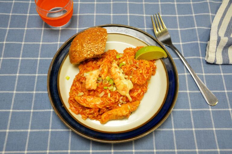 Jambalaya med rød tomatris, kyllingestrimler og jomfruhummerhaler anrettet på en hvid tallerken med en limebåd, hakket forårsløg og en sesambolle