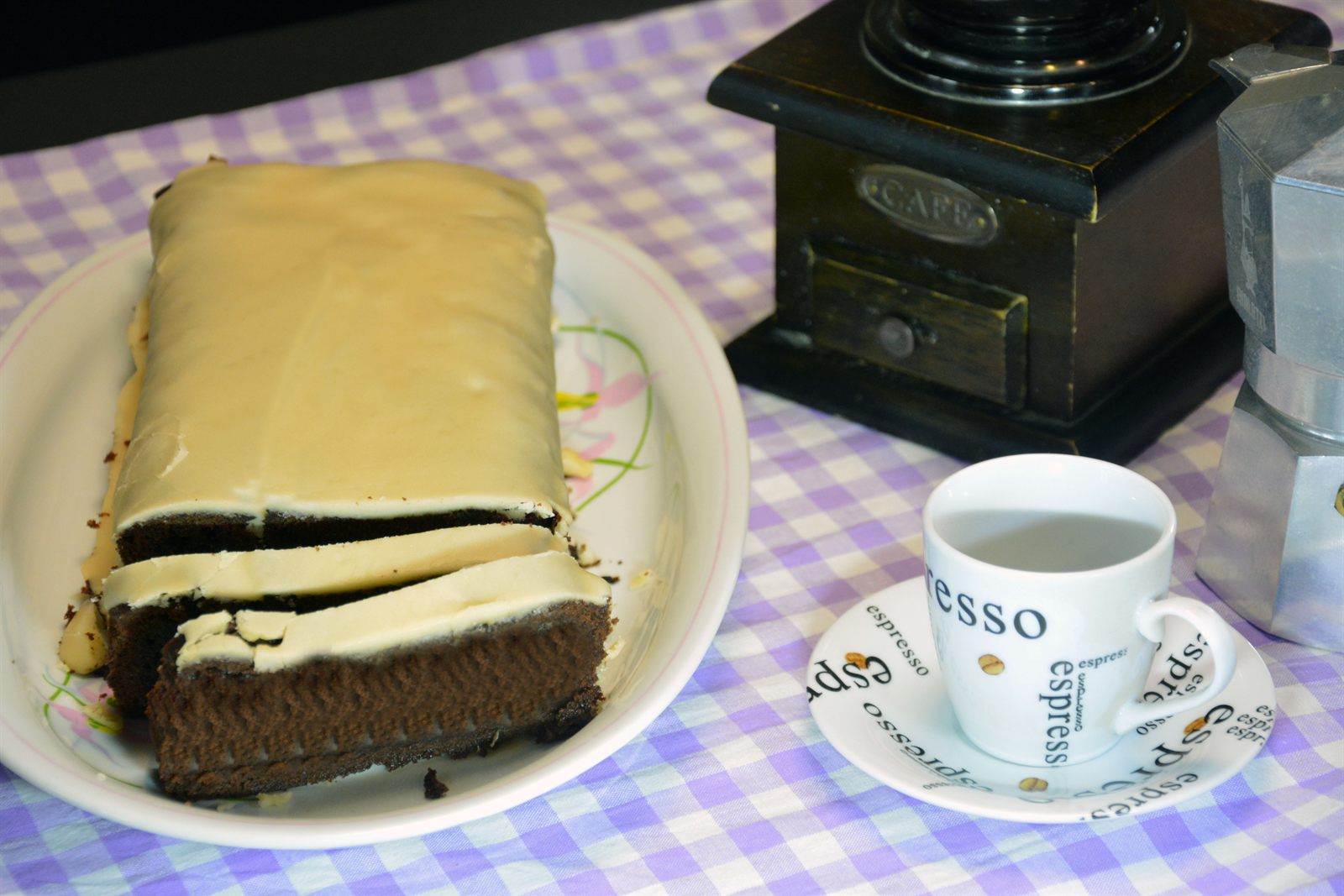 Kaffekage med lys kaffeglasur skåret i skiver på et hvidt fad, med espressokop, gammel kaffekværn og mokkakande ved siden af