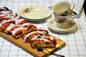 Klippet kanelstang med striber af hvid glasur på et aflangt træbræt, delvis skåret i stykker med kanelfyld i snitfladen, blomstrede kaffekopper og tallerkener på en blåternet dug bagved