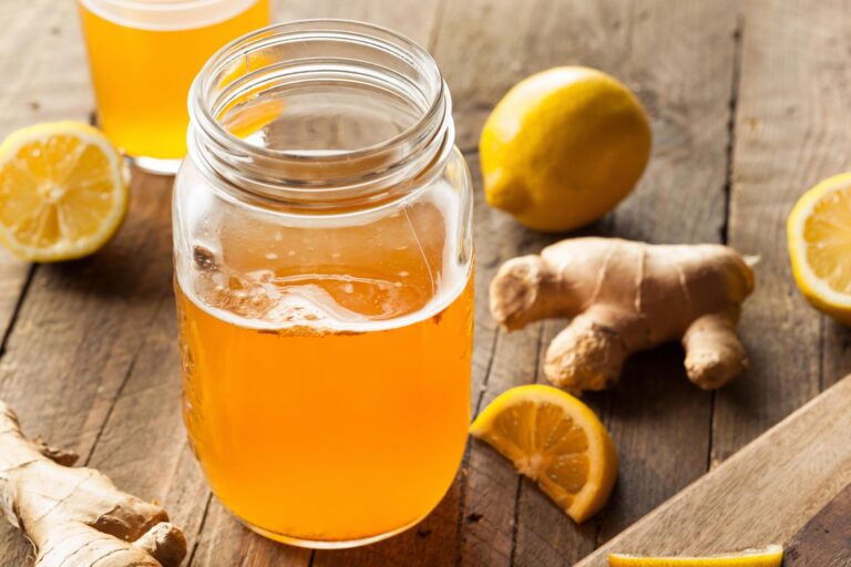 Hjemmelavet kombucha i et åbent glas på et træbord med friske ingefærrødder og halverede citroner, et glas kombucha i baggrunden