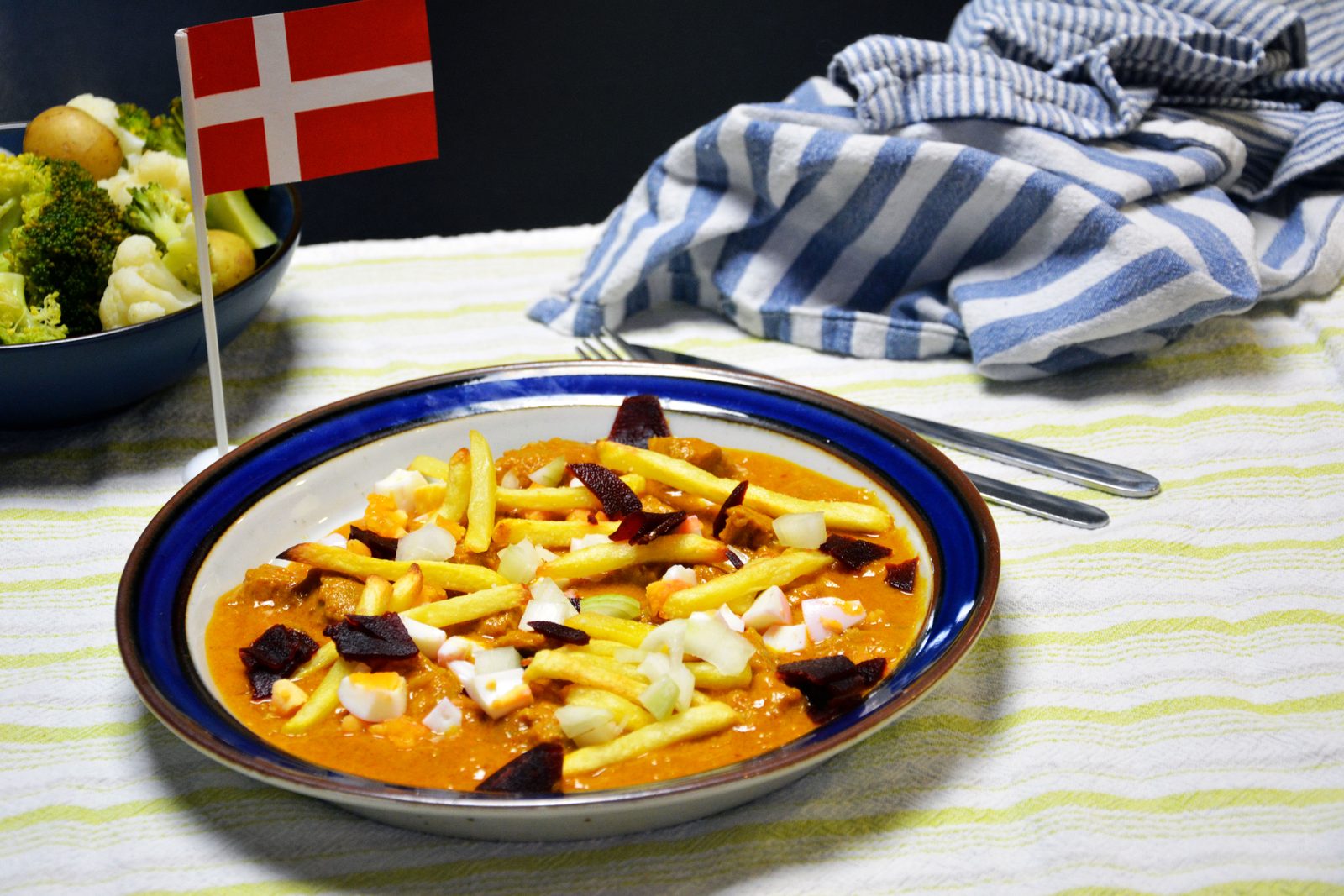 Kong Fiddes livret på en dyb tallerken med blå kant: kød i orange flødesovs med pommes frites, hakkede æg, løg og syltede rødbeder, et dannebrogsflag og en skål med kartofler, broccoli og blomkål i baggrunden