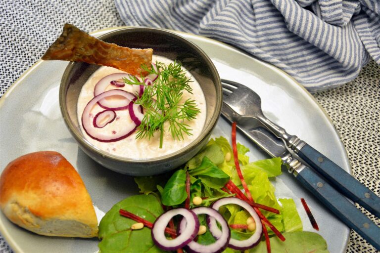Laksemousse i en grå skål pyntet med rødløgsringe, frisk dild og en stribe sprødt lakseskind, serveret med et stykke brød og grøn salat med rødbedestrimler og pinjekerner