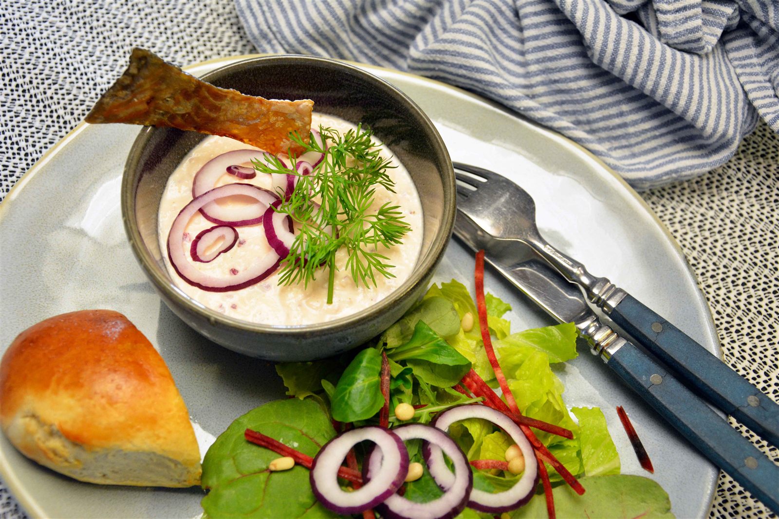 Laksemousse i en grå skål pyntet med rødløgsringe, frisk dild og en stribe sprødt lakseskind, serveret med et stykke brød og grøn salat med rødbedestrimler og pinjekerner