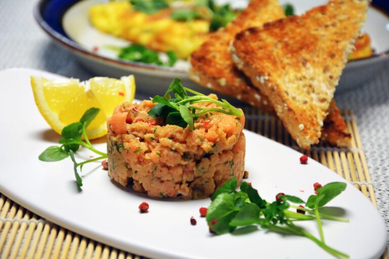 Et rundt tårn laksetatar med dild og rosa peberkorn, toppet med brøndkarse, på en aflang hvid tallerken med en citronbåd og løse rosa peberkorn. Bagved trekanter af ristet kernebrød og en tallerken med omelet