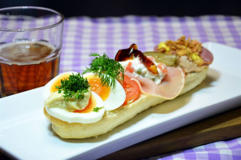 Et landgangsbrød på et langt hvidt fad med kogte æg, mayonnaise og karse, tomatskiver med dild, hamburgerryg med italiensk salat og sky samt leverpostej og spegepølse med ristede løg. Ved siden af står et glas øl på en lilla-ternet dug