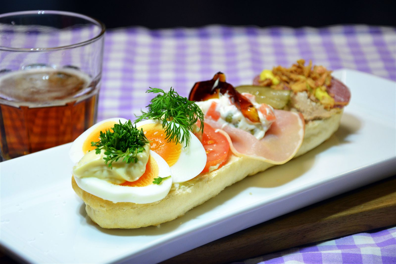 Et landgangsbrød på et langt hvidt fad med kogte æg, mayonnaise og karse, tomatskiver med dild, hamburgerryg med italiensk salat og sky samt leverpostej og spegepølse med ristede løg. Ved siden af står et glas øl på en lilla-ternet dug