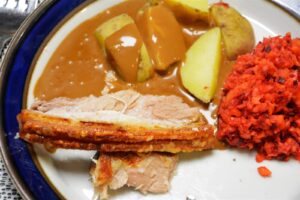 To skiver langtidsstegt ribbensteg med gyldne, sprøde svær på en tallerken med blå kant sammen med kogte kartofler i brun sovs og en råkostsalat af rødbede og gulerod