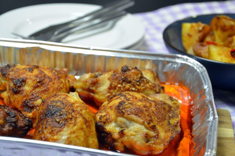 Fire gyldenbrune, paprikakrydrede kyllingeoverlår i en aluminiumsbakke med rødlig marinade i bunden, bagved en hvid tallerken med kniv og gaffel og en mørkeblå skål med ovnbagte kartoffelstykker og chili på en lilla-ternet dug
