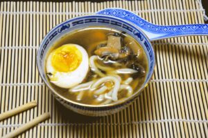 Miso suppe i en blåhvid porcelænsskål med udon-nudler, champignoner og et halvt æg, med en porcelænsske og spisepinde på en bambusmåtte